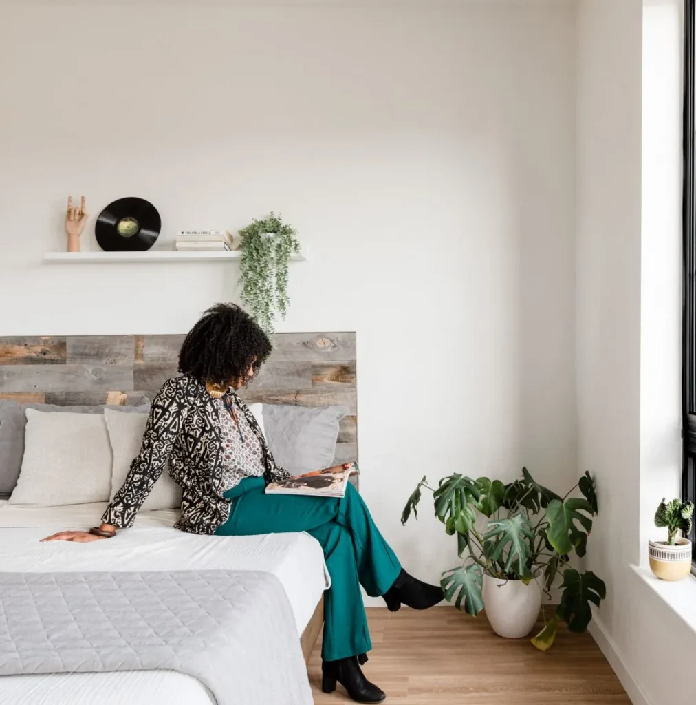 A person sits on a bed reading a magazine in a modern, minimal bedroom with plants and a shelf holding a vinyl record and decorative items.