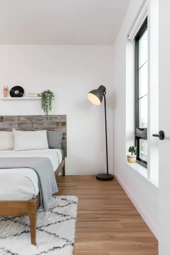 A modern bedroom with a wooden bed, white bedding, a gray blanket, a standing black floor lamp, a small plant by the window, and minimalist decor on a wall shelf.